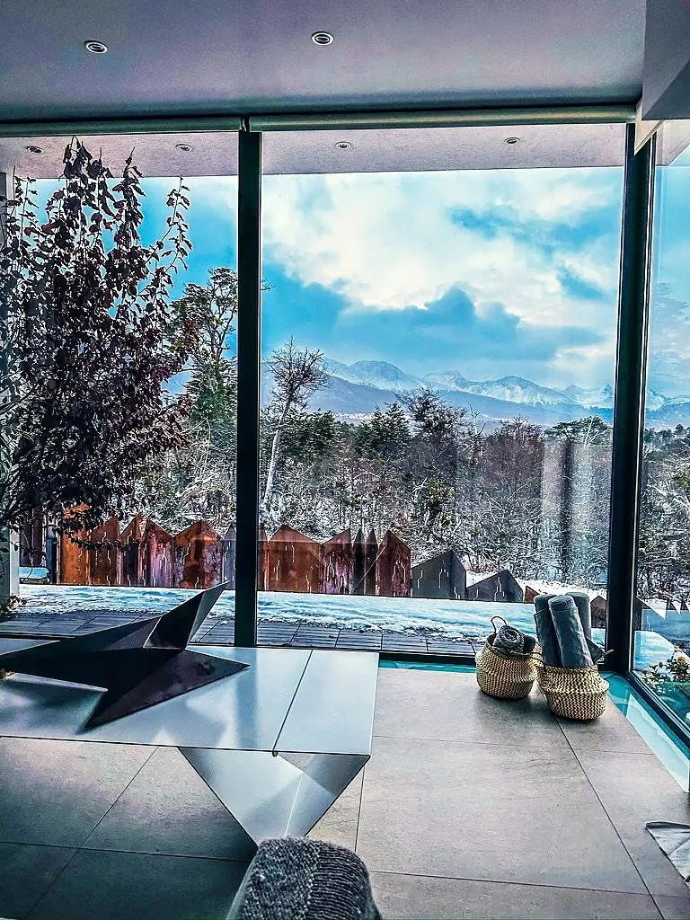 Estar con ventanal al bosque nevado, cerco de corten y mesa de acero plegado